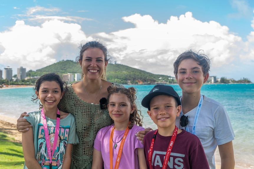 A mother and her four children, two sons and two daughters, smile for a picture with a beach and water in the background