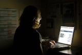 A woman sits at a desk working on her laptop in a dark room.