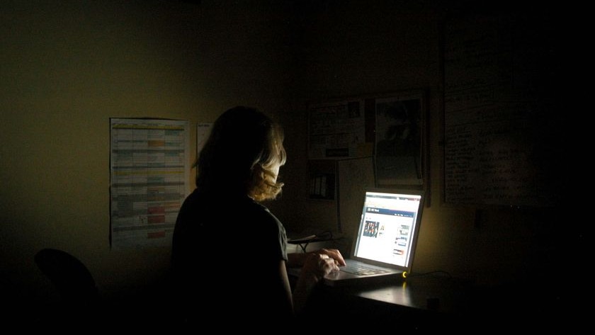 A woman sits at a desk working on her laptop in a dark room.