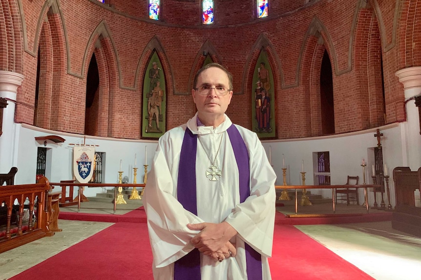 A bishop stands in a church looking at the camera with an altar in the background