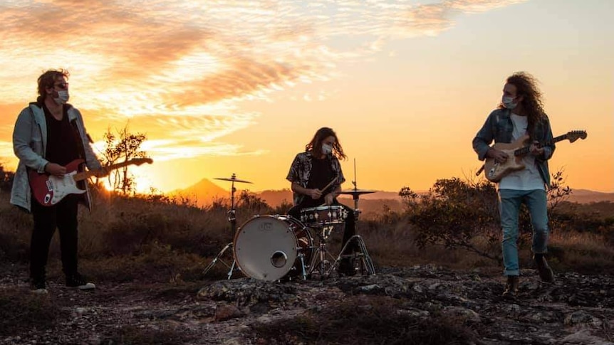 a three-piece band playing instruments outdoors in front of a sunset wearing surgical masks