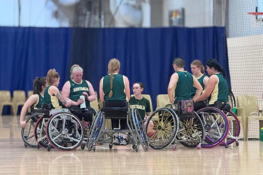 A group of Australian female wheelchair basketballers huddle around a coach who is giving instructions.