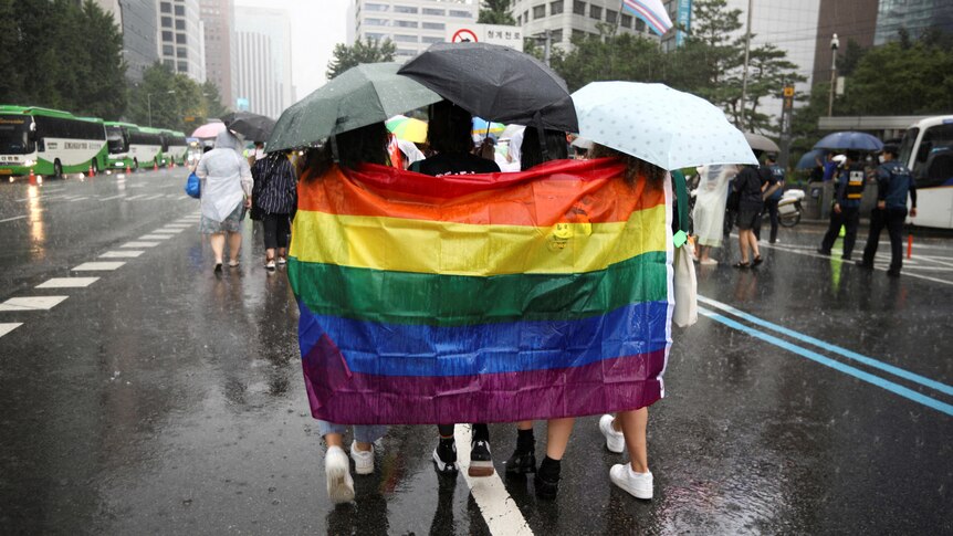 Three people walking holding a rainbow flag behind them