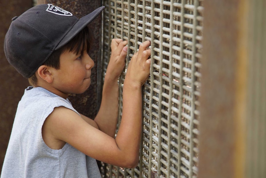 A young boy clings to the US-Mexico border fence as he talks to his grandfather on the other side.