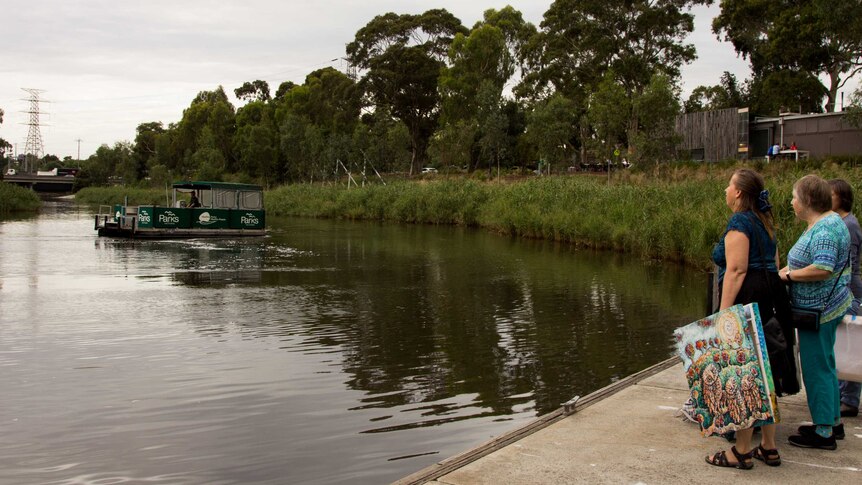 People holding artworks stand on a jetty by a river, watching a ferry punt come towards them.