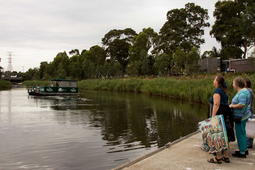 People holding artworks stand on a jetty by a river, watching a ferry punt come towards them.