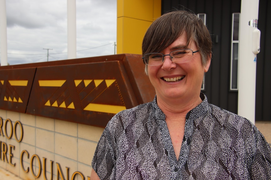 A woman with dark hair in a blue shirt smiles in front of a modern building.