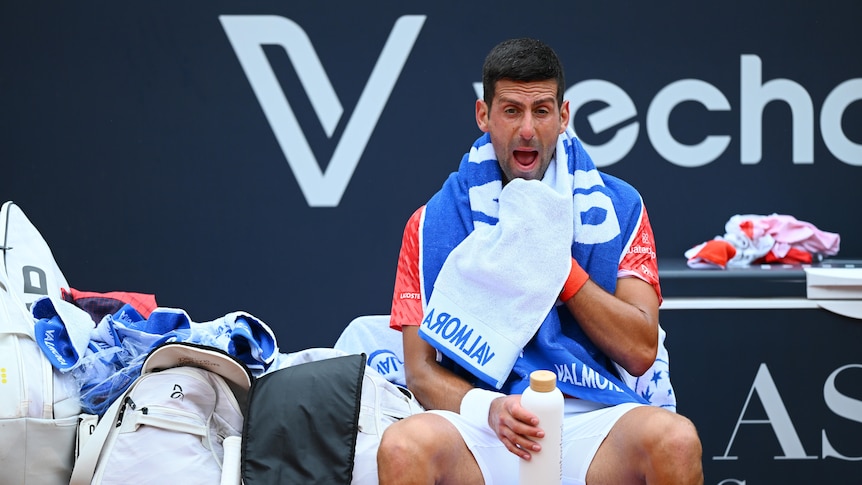 Tennis star Novak Djokovic sits at courtside holding a towel as he screams in frustration during a match.