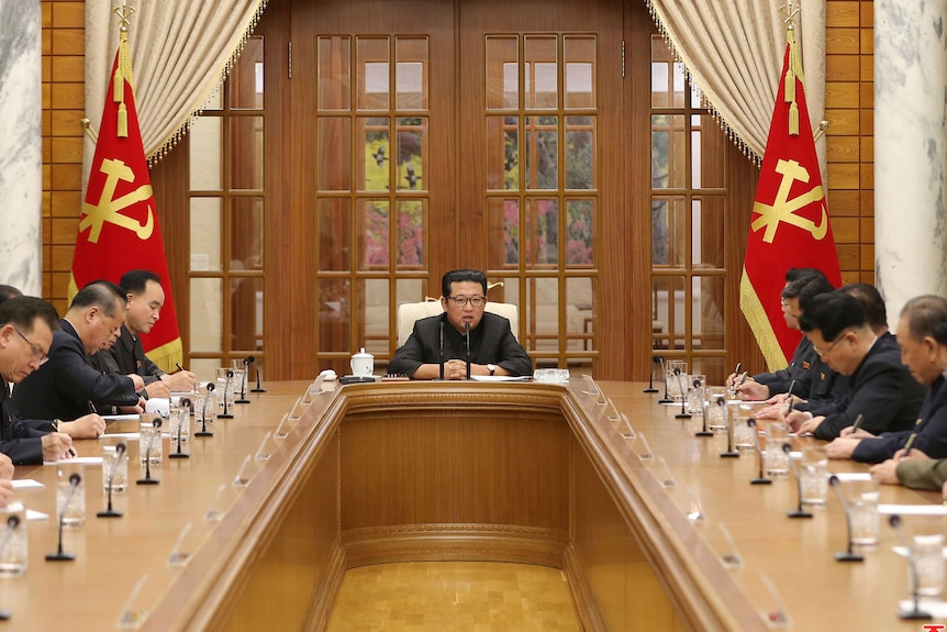 Korean officials line two sides of a meeting table, writing notes with leader Kim Jong Un speaking in the centre  