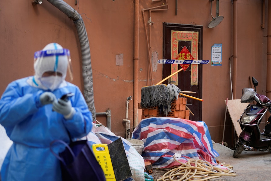 A person in full PPE stands outside an apartment which has a seal over the door.