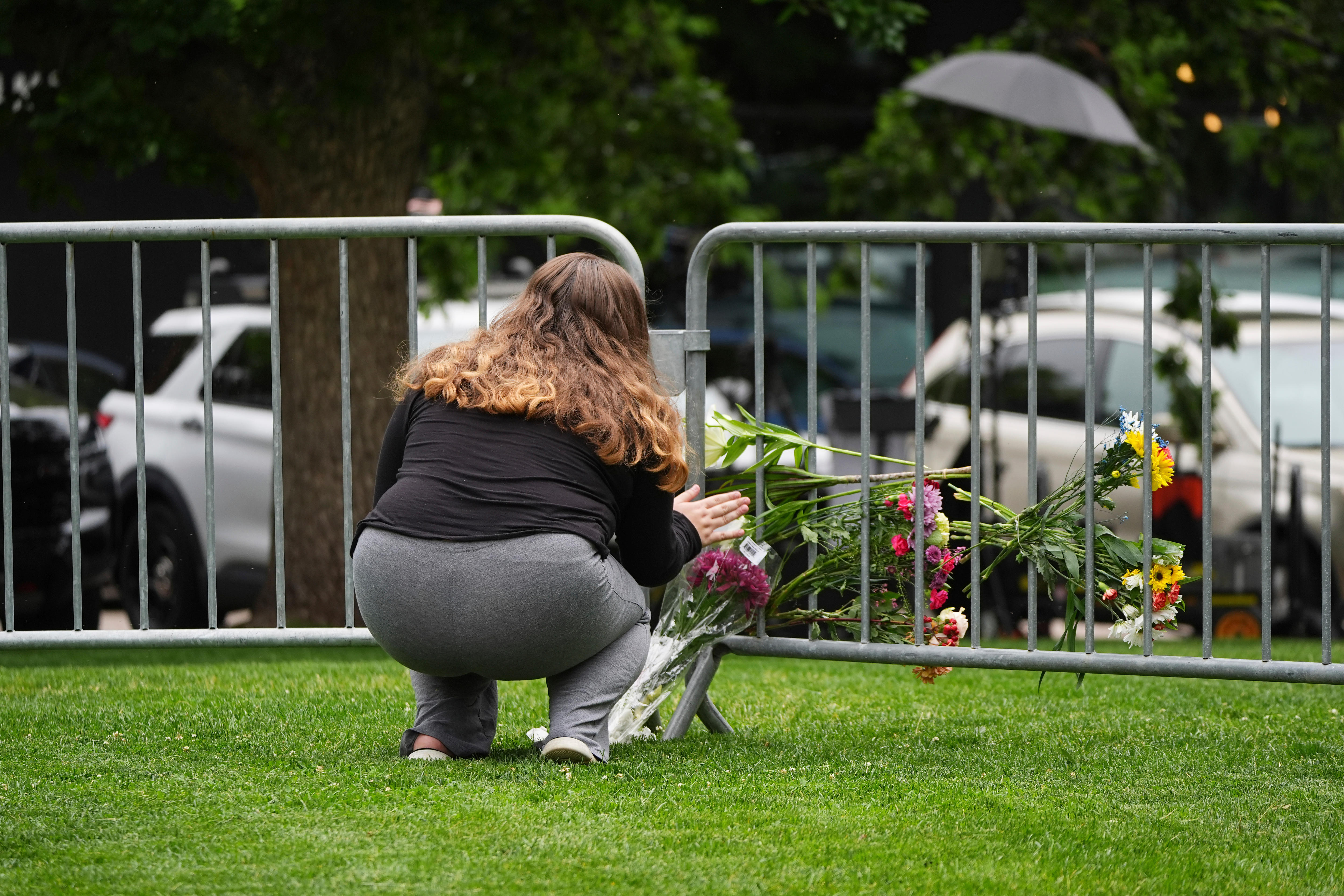 A brunette woman squatting on green grass alongside a fenceline dotted with flowers