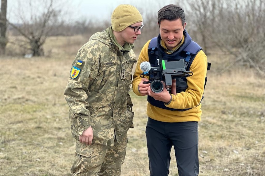 Man wearing bullet-proof vest showing a soldier what he is filming with his camera.