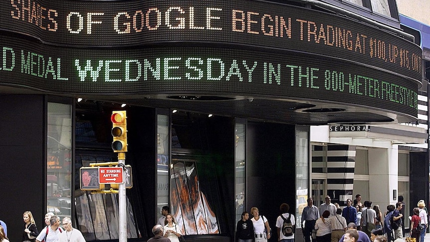 A neon strip of text above an office building with words 'shares of Google begin trading at $100'. Crowd of people walk below.