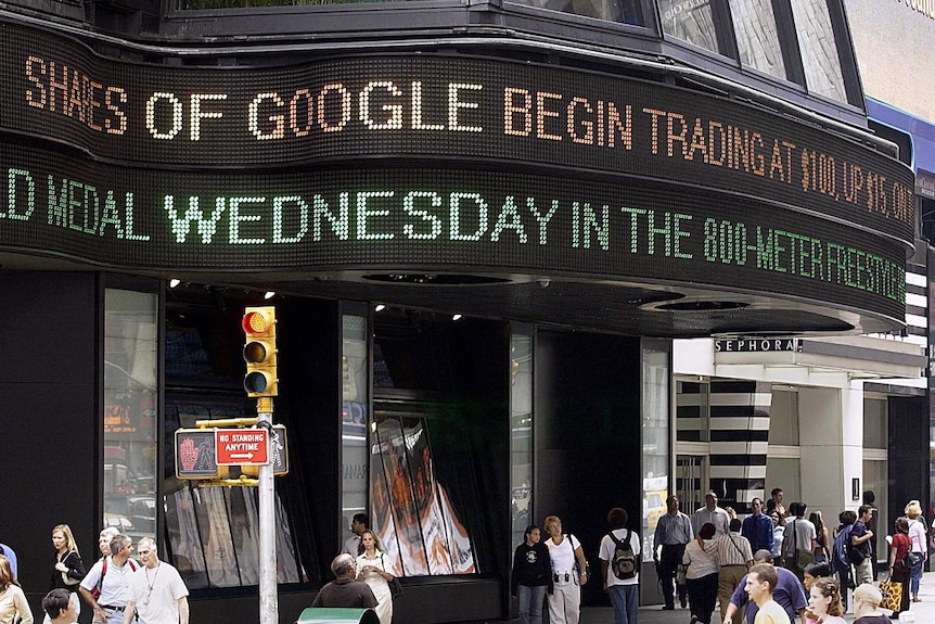 A neon strip of text above an office building with words 'shares of Google begin trading at $100'. Crowd of people walk below.