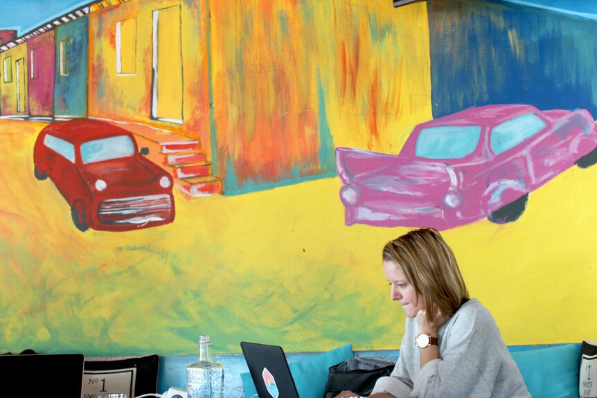 Colourful painting of two cars on a wall and a woman working on a computer below