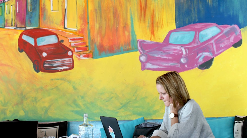A woman works at a laptop in a cafe. There is a colourful painting of two cars on the wall behind her