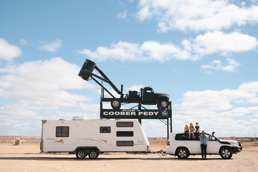 At a distance, a large white caravan attached to a white four-wheel drive, on red dirt before a large 'Coober Pedy' sign.