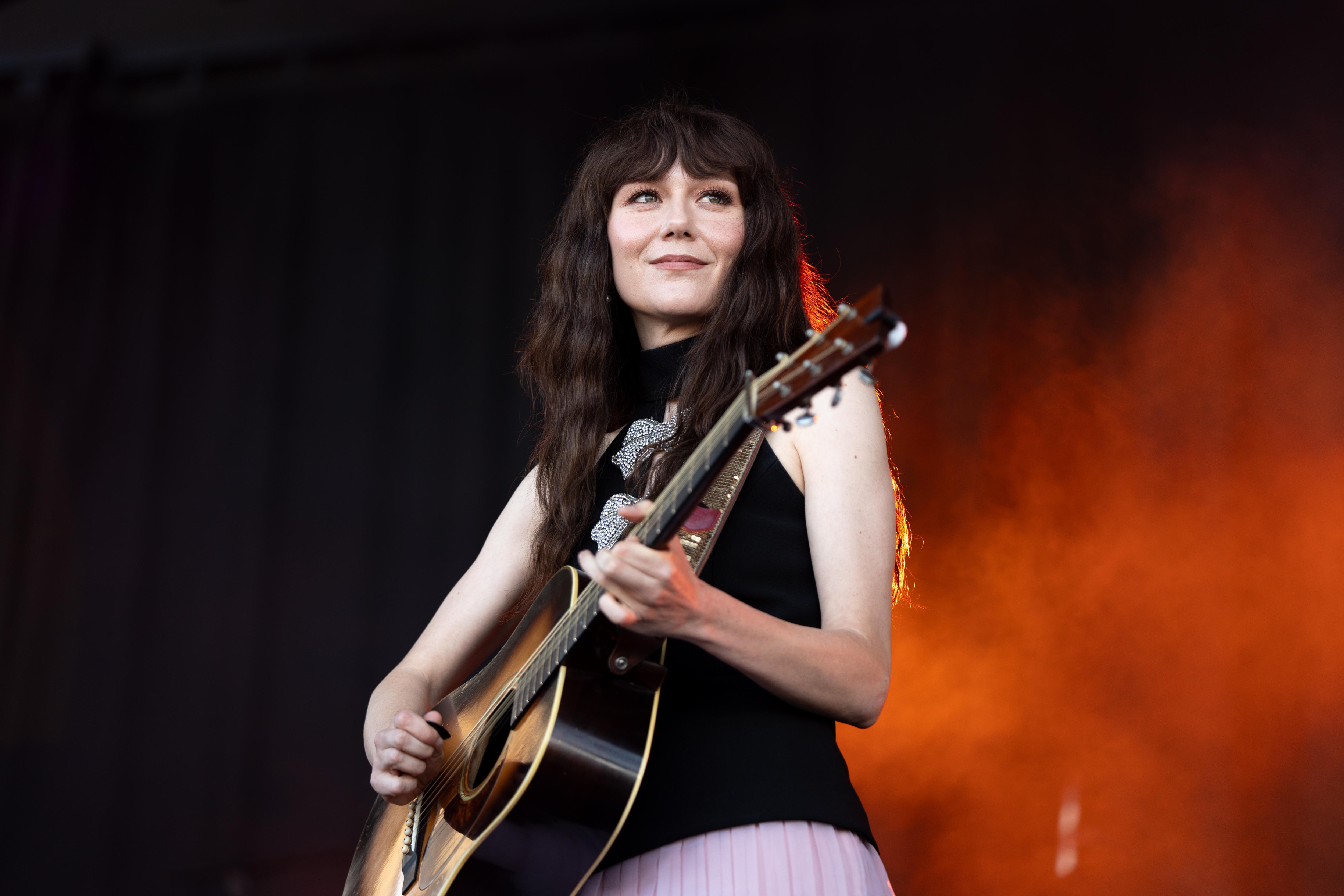 A woman with long dark hair and a fringe smiles on stage while holding her acostic guitar, mid-performance