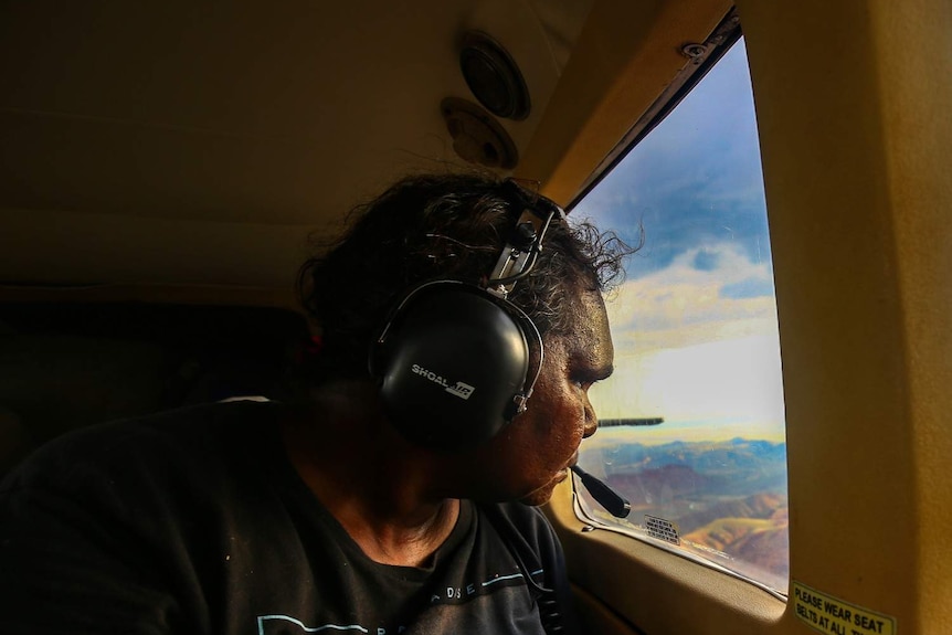 A woman wearing a black tshirt and a headset looks out a plane window.