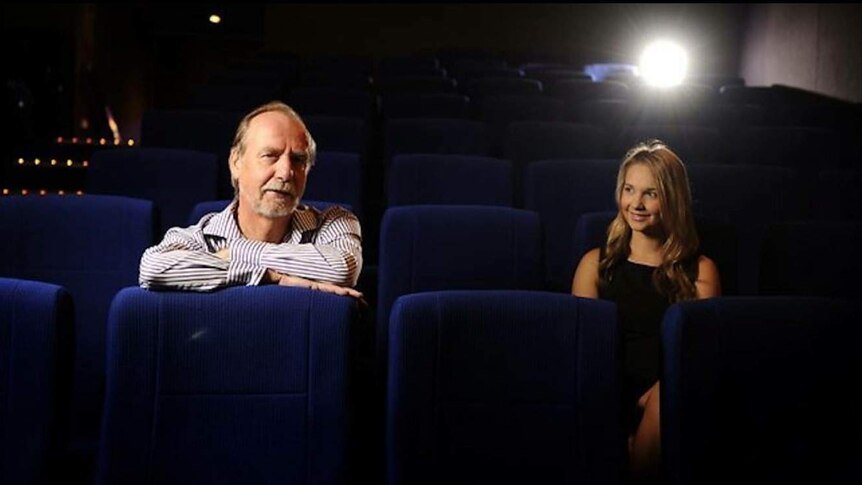 Tom and Ellie sit in cinema seats. He looks at the camera and she smiles, while a projection light can be seen.