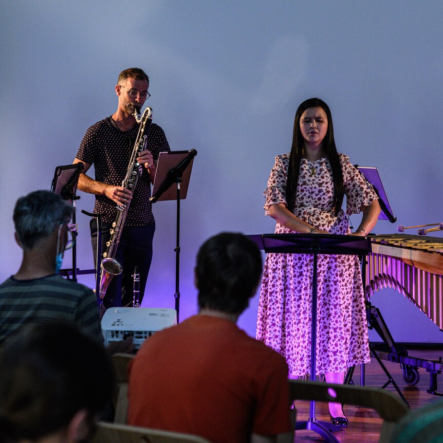 A flute player, bass clarinettist, singer reading braille music and vibraphone player perform on stage.