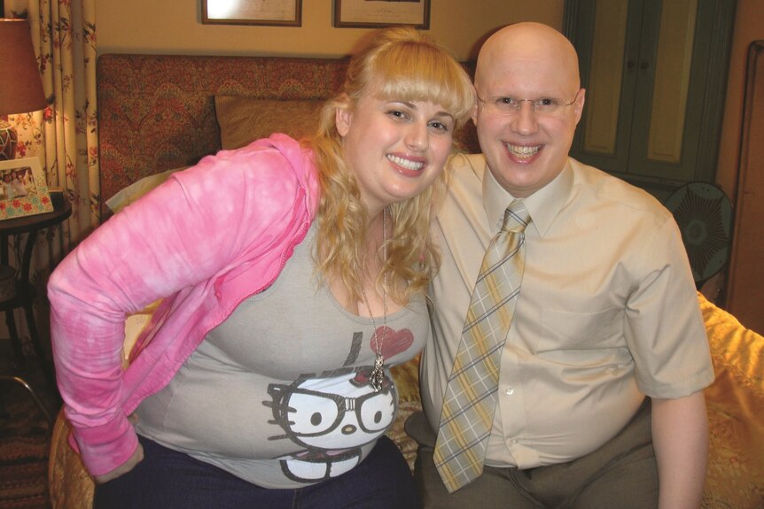 Rebel Wilson with long blonde hair in a pink cardigan and tan top with Matt Lucas wearing glasses, a shirt and tie
