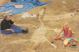 Two women sit in red dirt surrounded by tarps and boxes. Between them is a fossil of a large dinosaur. They hold tools.