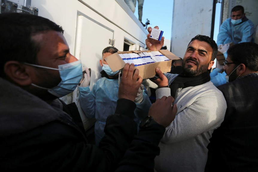 A man with a box of vaccine vials surrounded by people in face masks