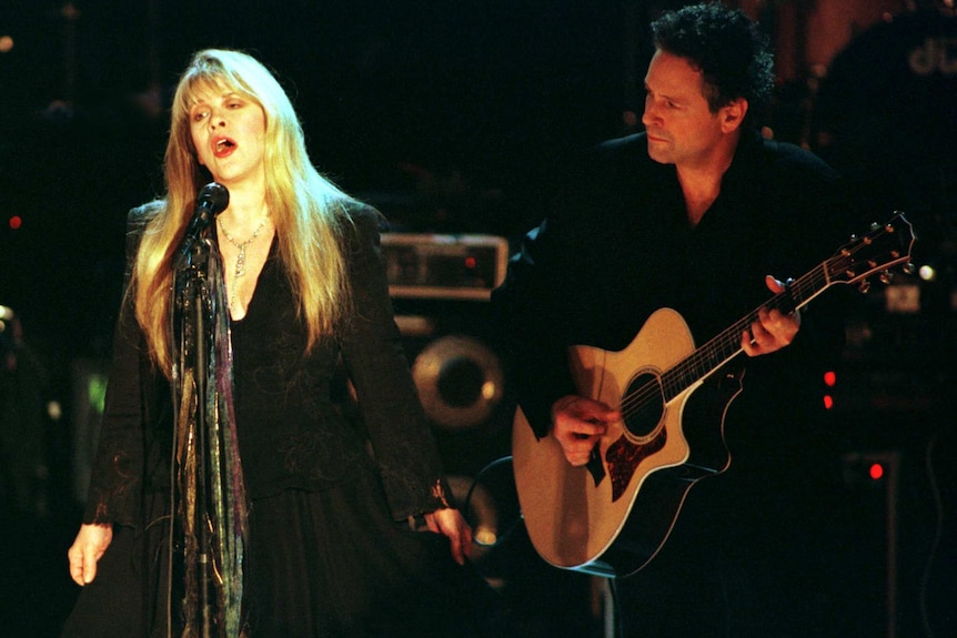 Stevie Nicks sings into a microphone while Lindsay Buckingham plays an acoustic guitar.