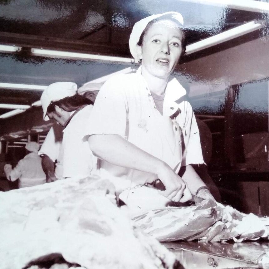 A woman in a white cap cuts into an animal carcass in an assembly line.