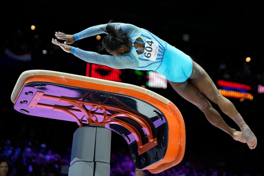 Simone in motion as she throws her body over a vault, her arms and legs stretched out as if she's diving.
