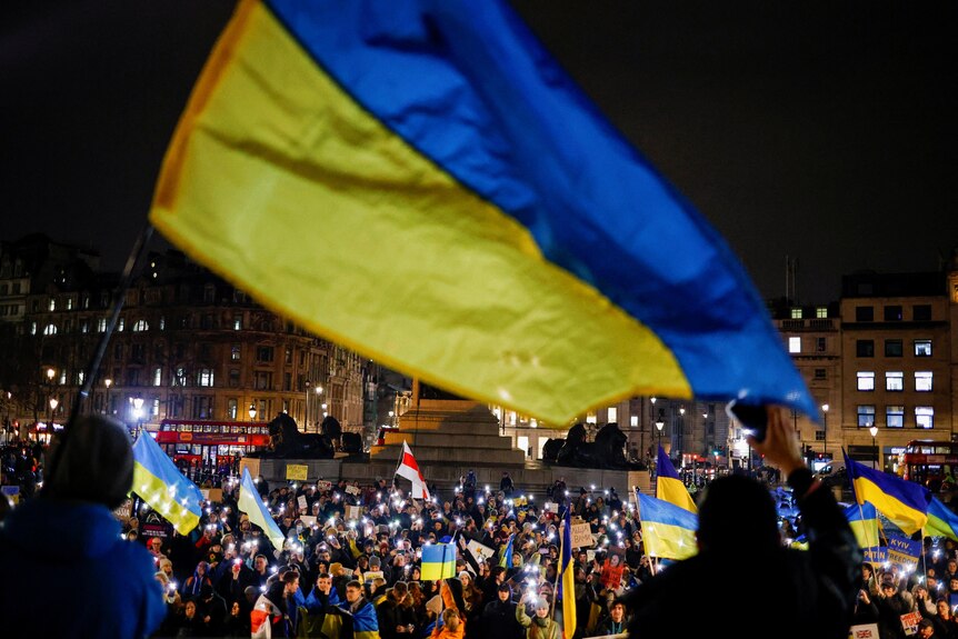 Large Ukraine flag flies at night time, crowds of people in background.