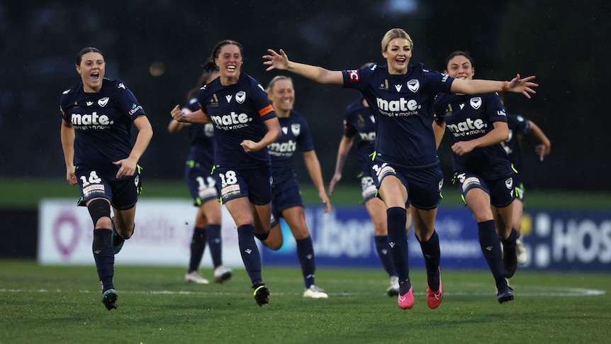 A team of soccer players wearing navy blue run towards the camera after winning a game