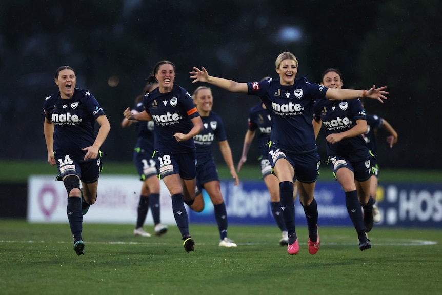 A team of soccer players wearing navy blue run towards the camera after winning a game