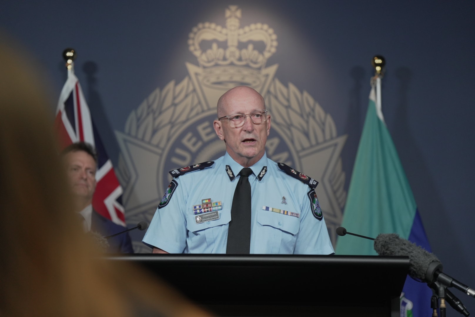 Um policial careca falando em um púlpito em frente ao logotipo do QPS.