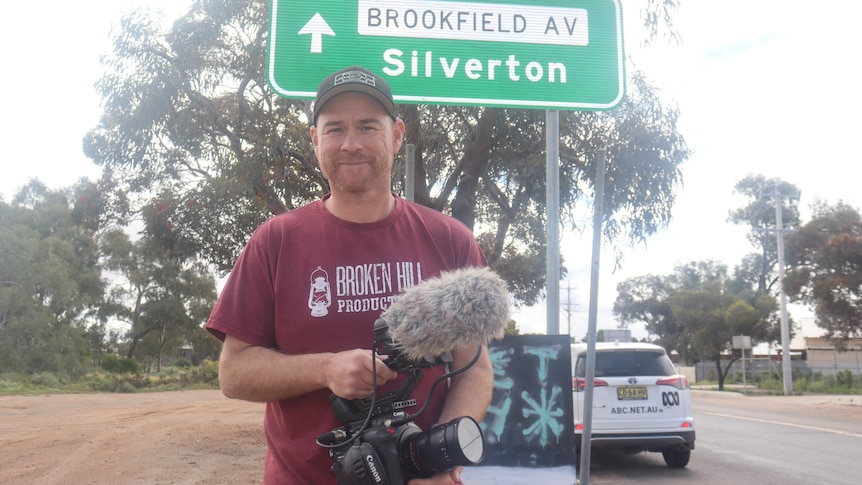 A man wearing a red shirt and a cap holding a camera, standing in front of a green sign that says 'Silverton' and a tree.