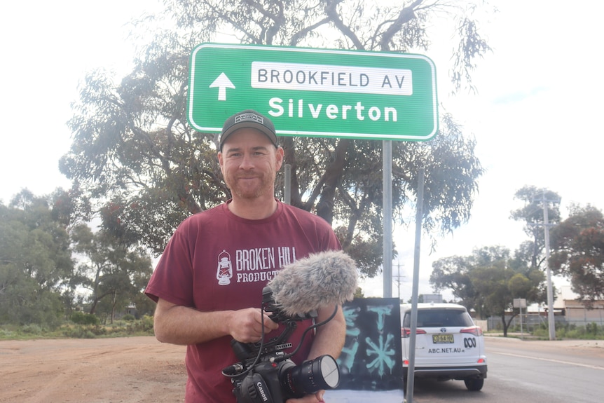 A man wearing a red shirt and a cap holding a camera, standing in front of a green sign that says 'Silverton' and a tree.