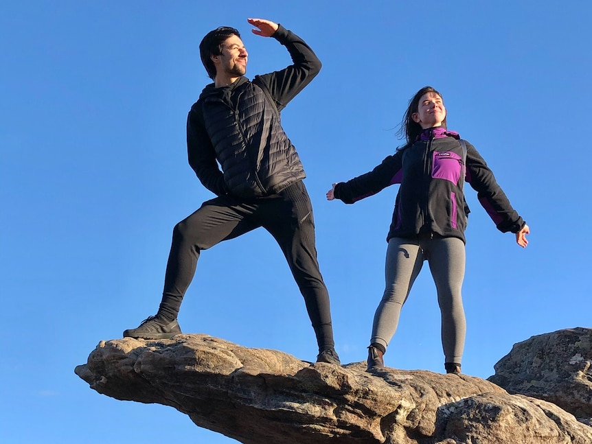 A man and a woman pose on a rocky cliff against a blue sky.