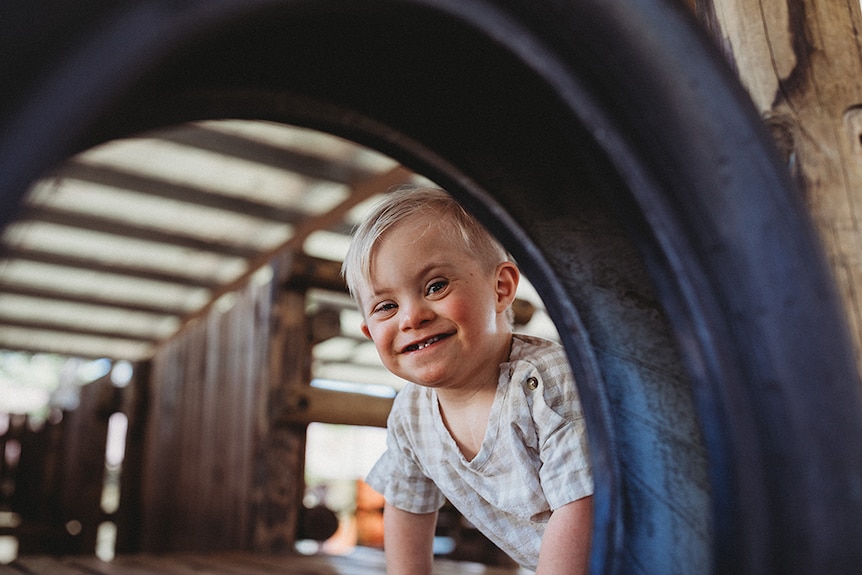 Grayson, a blonde boy with a cheeky smile, looks at the camera through a dark circular shape.