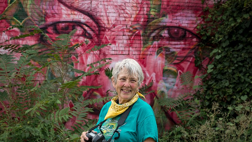 A woman smiles holding camera and standing in front of brightly decorated mural wall with overgrown garden.
