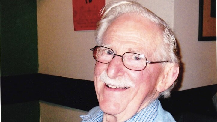 A man with white hair and moustache, and glasses, sits indoors smiling and holding a glass.