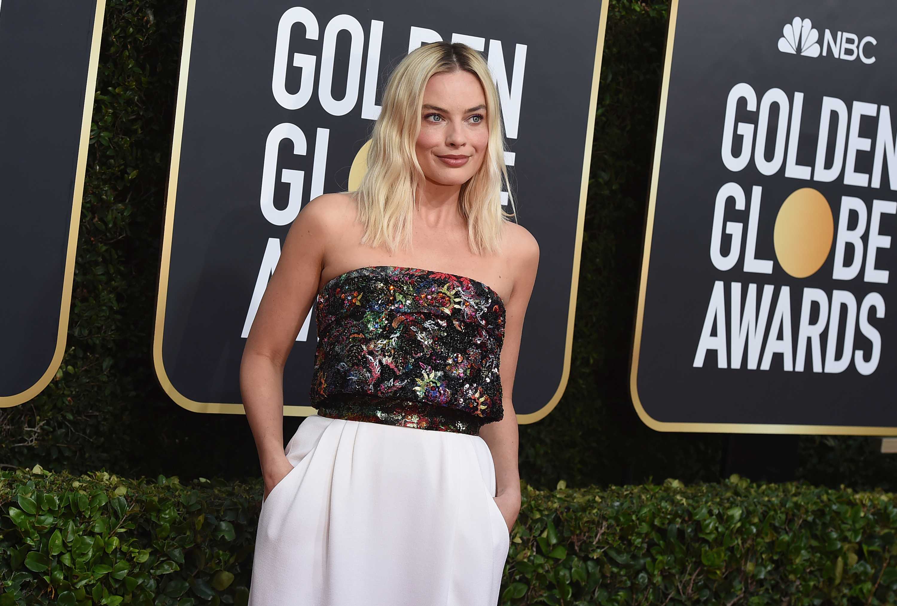 Margot Robbie smiles for the camera wearing a multi-coloured sleeveless top and a white floor-length skirt.