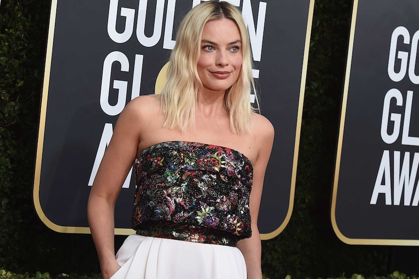 Margot Robbie smiles for the camera wearing a multi-coloured sleeveless top and a white floor-length skirt.