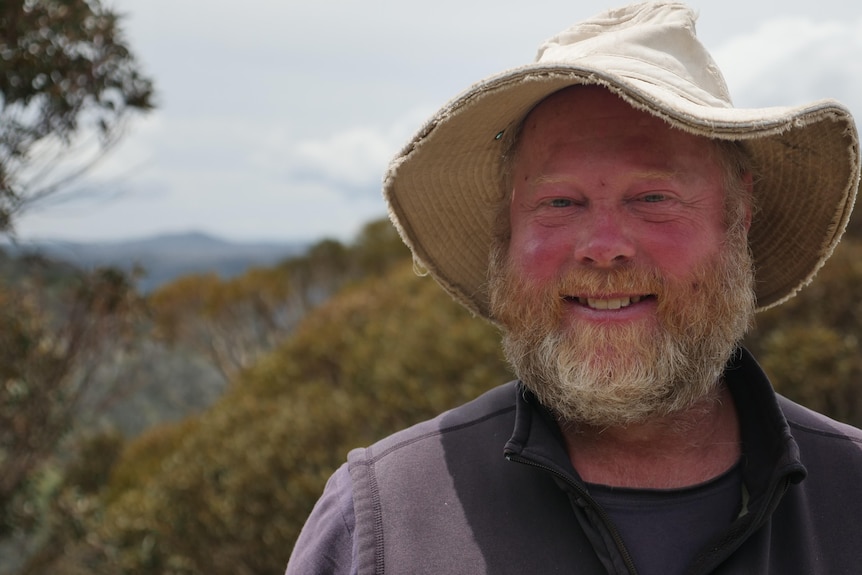 A man in a hat smiles at the camera while Victoria's High Country peaks and trees stretch into the background
