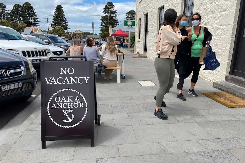 People walk past a 'no vacancy' sign outside a hotel restaurant on a sunny day.