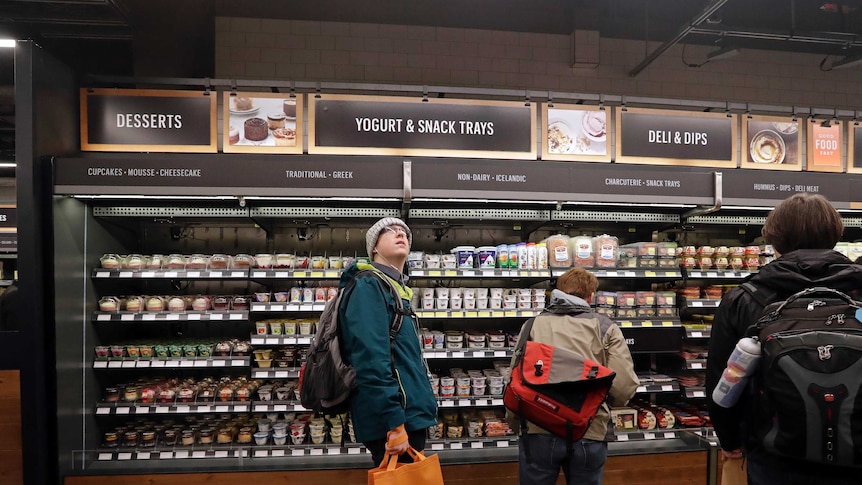 A customer looks overhead in an Amazon Go store, in front of rows of desserts and yoghurts.