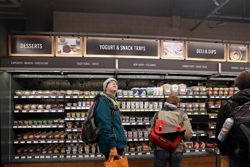 A customer looks overhead in an Amazon Go store, in front of rows of desserts and yoghurts.