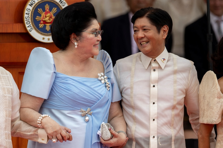 Imelda Marcos in a sky blue dress with her trademark beehive hairdo stands next to her son Bongbong as they smile at each other