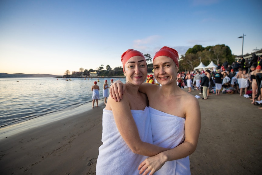 Two women stand in towels and red swimming caps at the beach with their arms around each other.