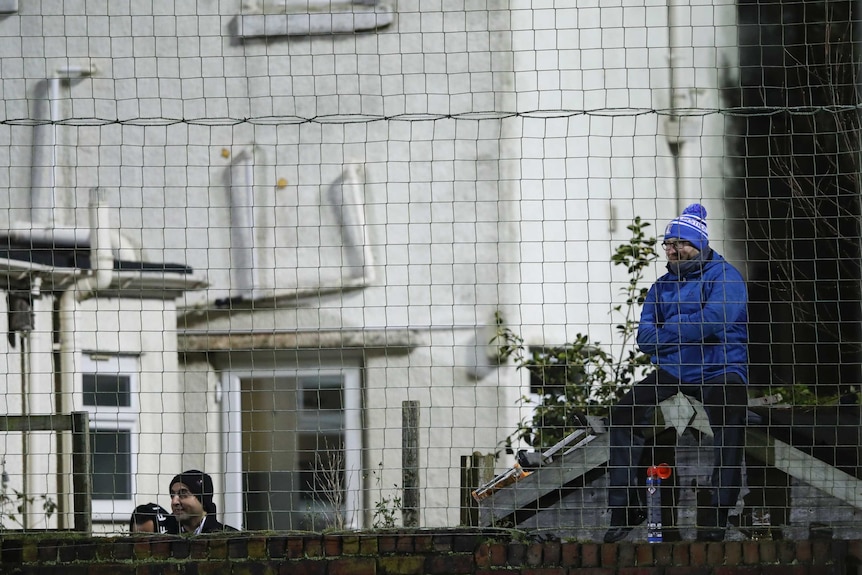 A man wearing a blue coat and bobble hat sits on a shed, with two others standing to his right